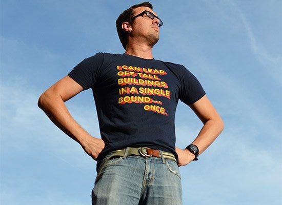 White man in black-framed glasses wearing jeans and a navy t-shirt with the text "I can leap buildings in a single bound... once" with blue sky behind