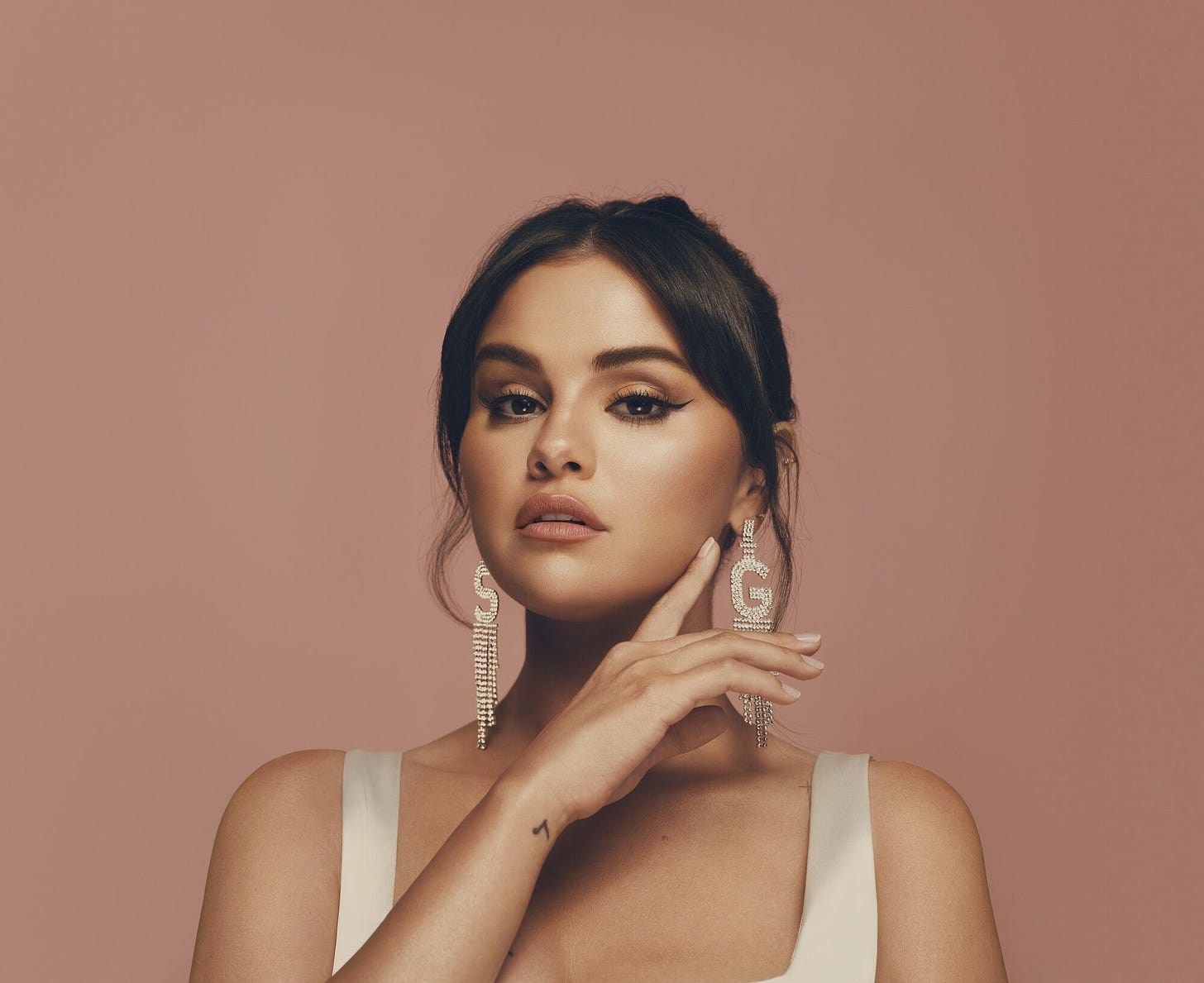 Selena Gomez poses with her hand under her chin, wearing long dangly earrings in the shape of an S and G, in front of a brown background.