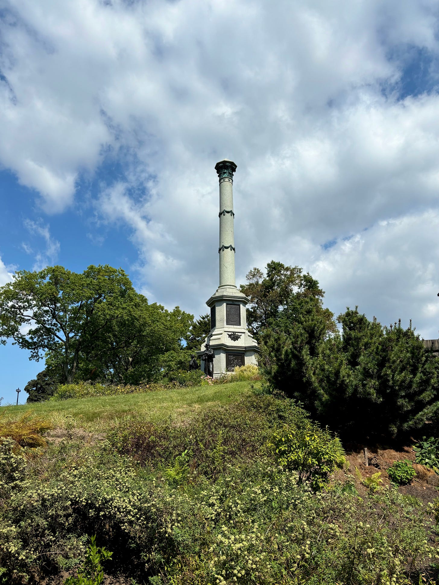 Looking up at the Battle Hill monument from the base of the hill. It is a column engraved with metal wreaths and stars atop a square base with statues of soldiers at each corner. The sky is blue and full of fluffy clouds and the flora is green in the heat of late summer. 