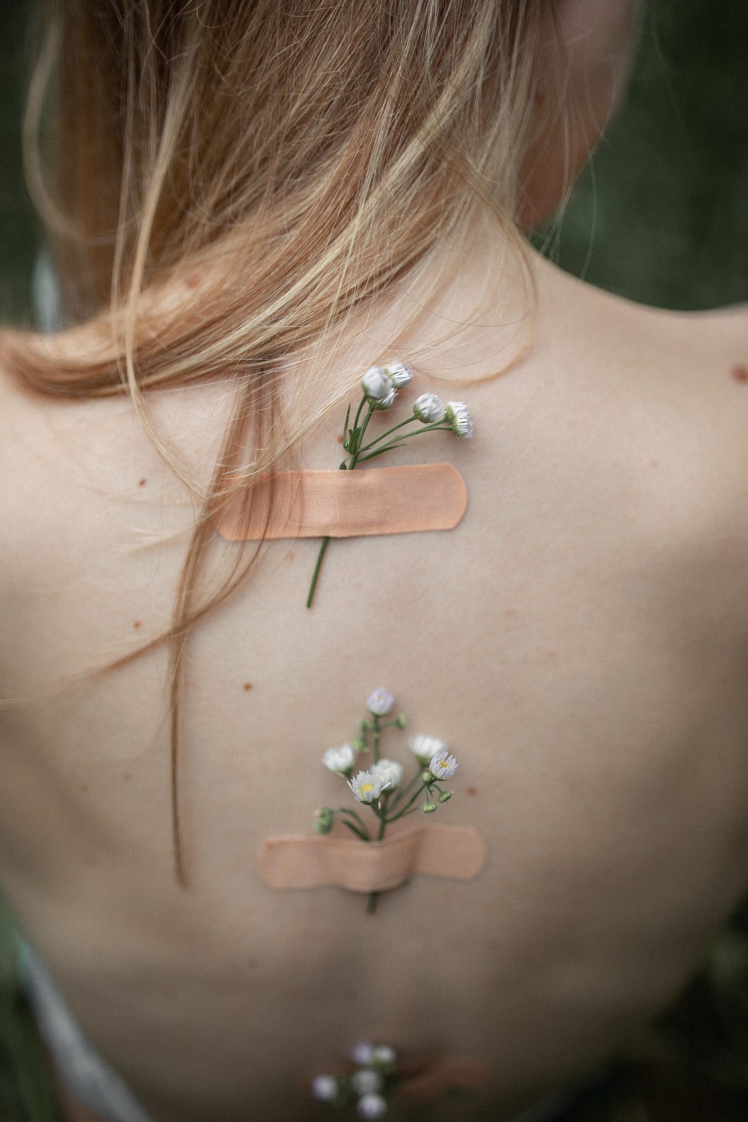 bare backed woman with long hair and three bandaids down her spine. Each bandaid has a sprig of flowers underneath.