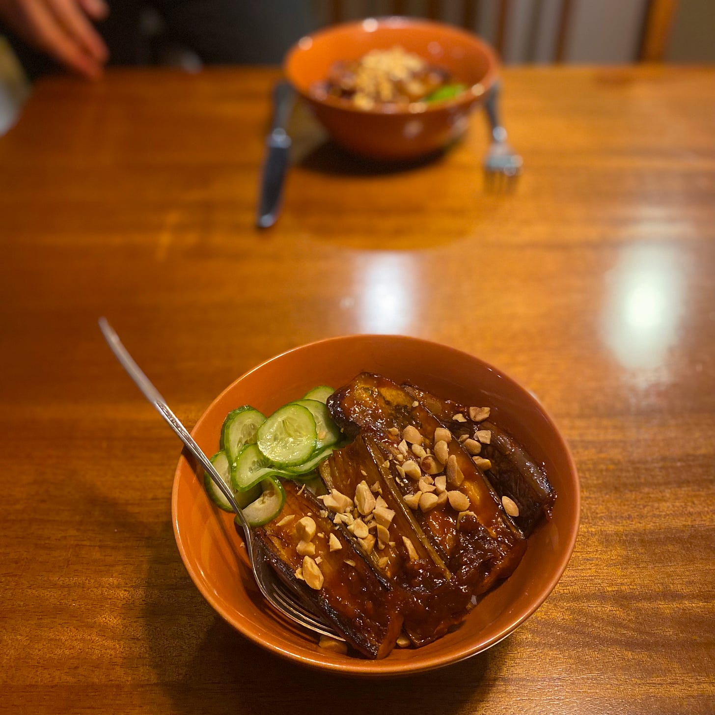 An orange bowl of gochujang-glazed eggplant with thinly sliced cucumber pickle on the side, and chopped peanuts sprinkled over the top.