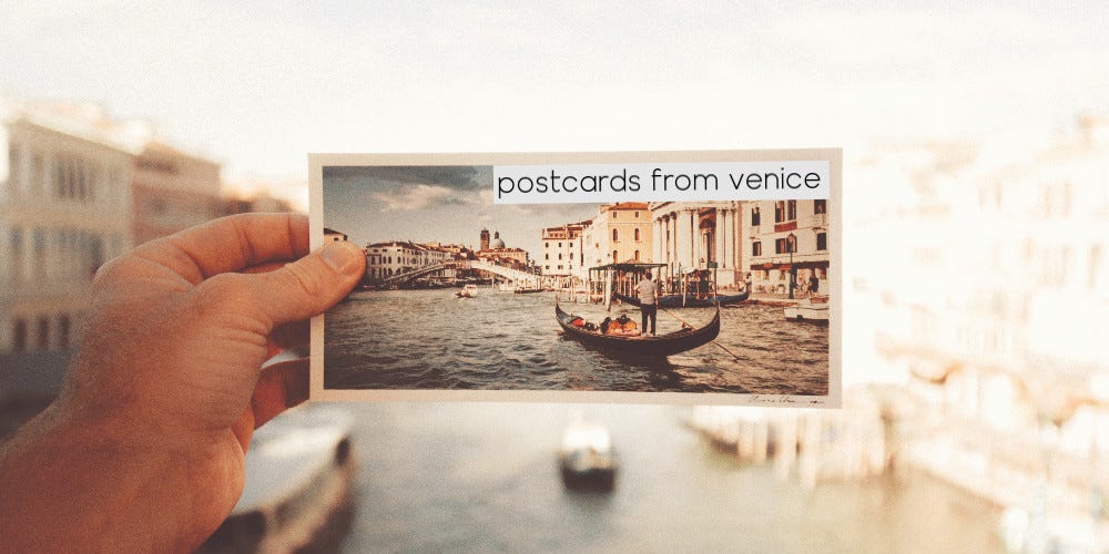 A man's hand holds a postcard of Venice in front of its real-life location on Grand Canal, Venice