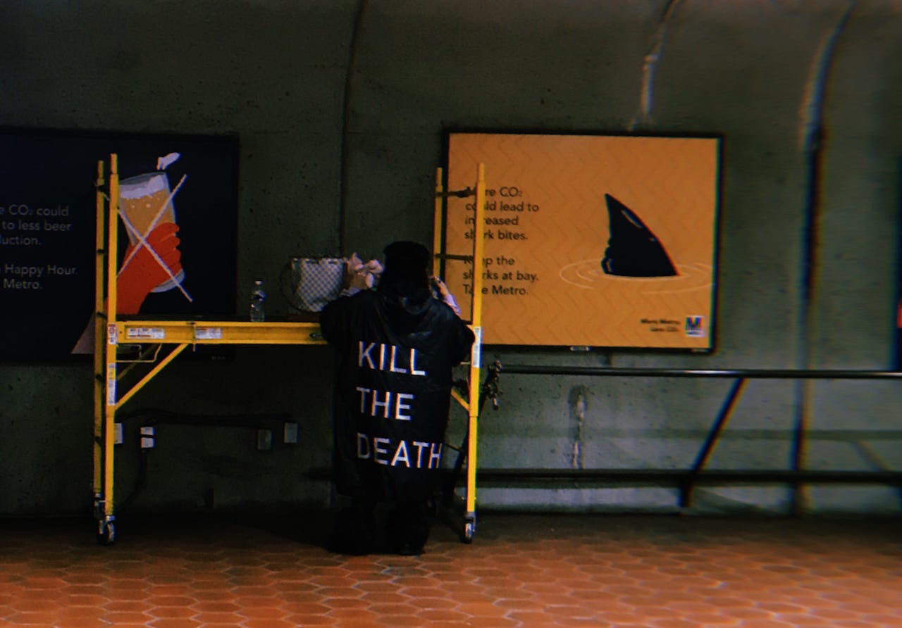 A dim, grainy photo of a small woman with her back to the camera, standing inside a metro station. The woman is rummaging through her handbag, which she has set on a yellow construction scaffolding. She is wearing an immense black coat that reaches nearly to the ground. On the back is embroidered “KILL THE DEATH” in all capital letters.