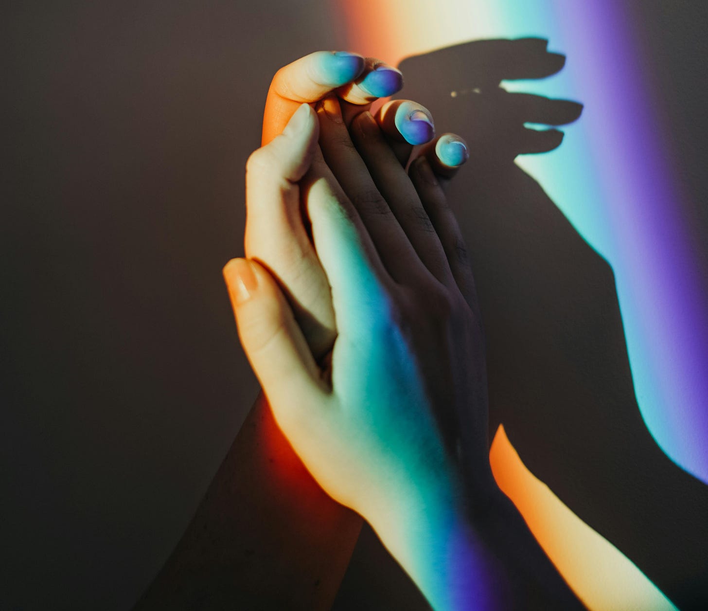Two hands holding each other. A rainbow of light is projected over them.