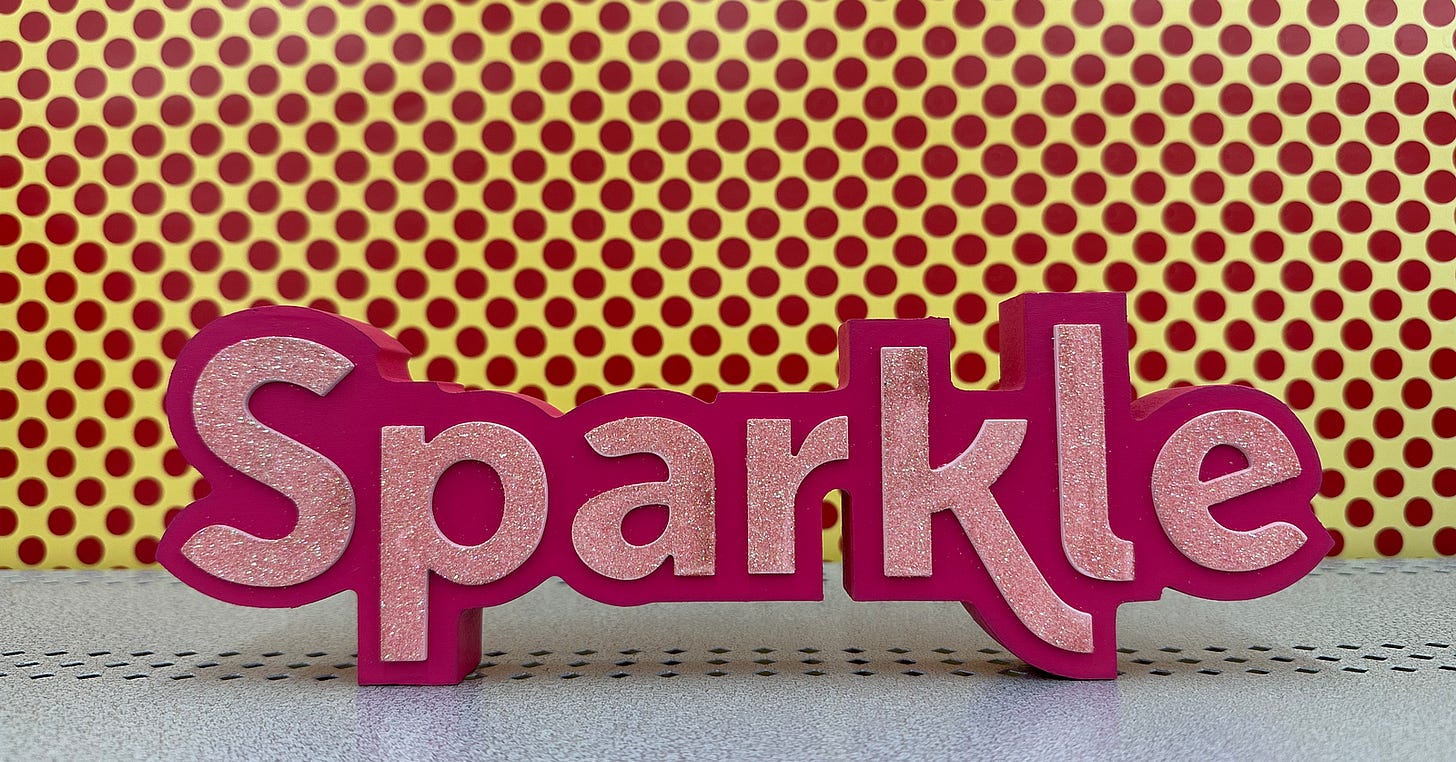 Pink sign that says “Sparkle” against a yellow backdrop with pink polkadots.