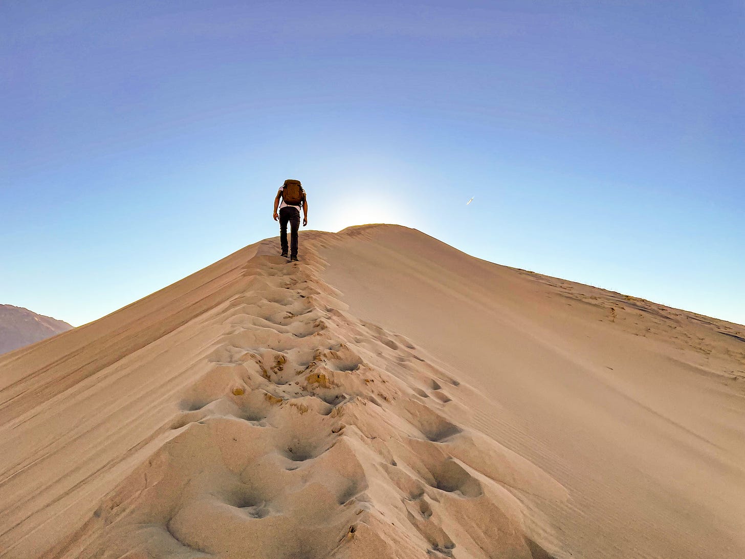 A man hikes up a sand dune that rises so high it blocks out the sun on a cloudless day.