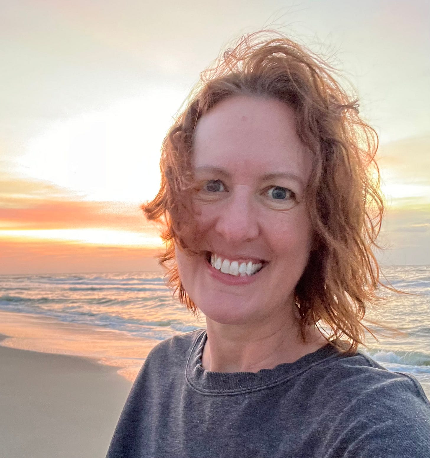 headshot of Marla, a white woman with short wavy hair, smiling at the beach with the ocean at sunset behind her