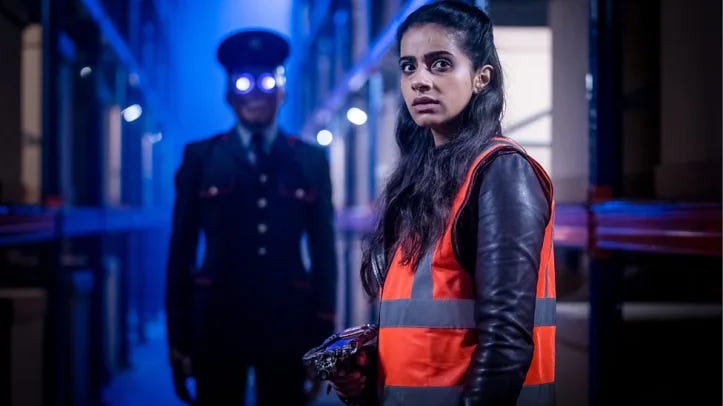 This image shows Yasmin "Yaz" Khan (played by Mandip Gill) standing in a Kerb!am Corporation warehouse corridor wearing a worker's uniform and holding a small inventory device. A Kerb!am Corporation delivery bot, with its eyes glowing blue, stands behind Yaz. This image shows Yasmin "Yaz" Khan (played by Mandip Gill) standing in a Kerb!am Corporation warehouse corridor wearing a worker's uniform and holding a small inventory device. A Kerb!am Corporation delivery bot, with its eyes glowing blue, stands behind Yaz.