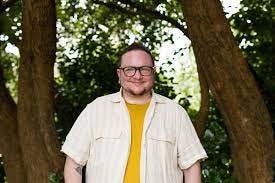 Rabbi Kukla stands in front of green bushes and a tree. They have white skin, a beard and wear glasses