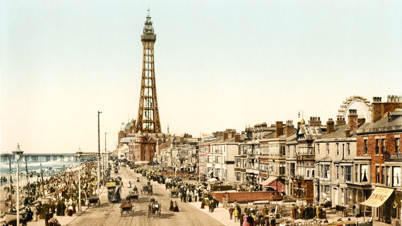 Das Bild zeigt ein altes, coloriertes Foto von der Strandpromenade in Blackpool. Zentral kann man auf den Aussichtsturm Blackpool Tower sehen. Links davon ist ein Stück Meer mit einem der Piers zu sehen, rechts vom Tower schaut ein Riesenrad über den Hausdächern hervor. Die Promenade ist voller Menschen, die spazieren gehen oder in Pferdekutschen unterwegs sind.