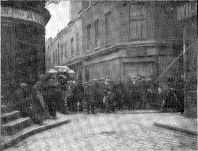 Murky black and white long-shot taken during shooting of early Alfred Hitchcock film Mrs Peabody (Number 13) in Limehouse, London. A crowd is visible in the cobbled street