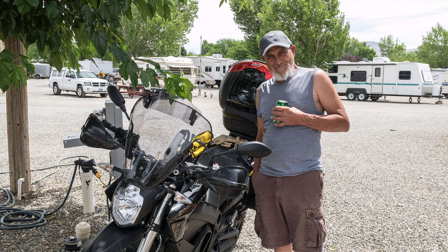 A man poses besides the Zero electric motorcycle as it charges at the RV park. He is wearing cargo shorts, a grey tank top, and a baseball cap. He has a white beard and is holding a green soda can in his left hand as he smiles and looks at the camera. Behind the motorcycle is the RV electrical panel that the bike is connected to. Beyond is the gravel parking area of the RV park with modest white travel trailers and a white pickup truck in the background.