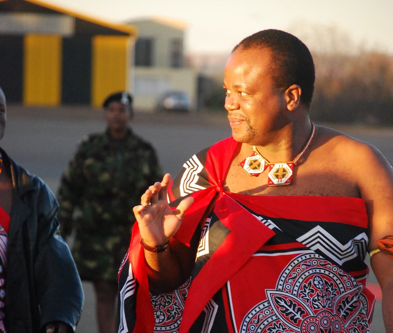 "File:Mswati III King of Eswatini.jpg" by Kollmeierf is licensed under CC BY-SA 3.0. Image shows the King in traditional dress and accompanied by a member of the armed forces in uniform in the background. The King is a middle-aged Black man with short cropped hair and a slight amount of facial hair. He is smiling and standing in sunlight from a setting sun.