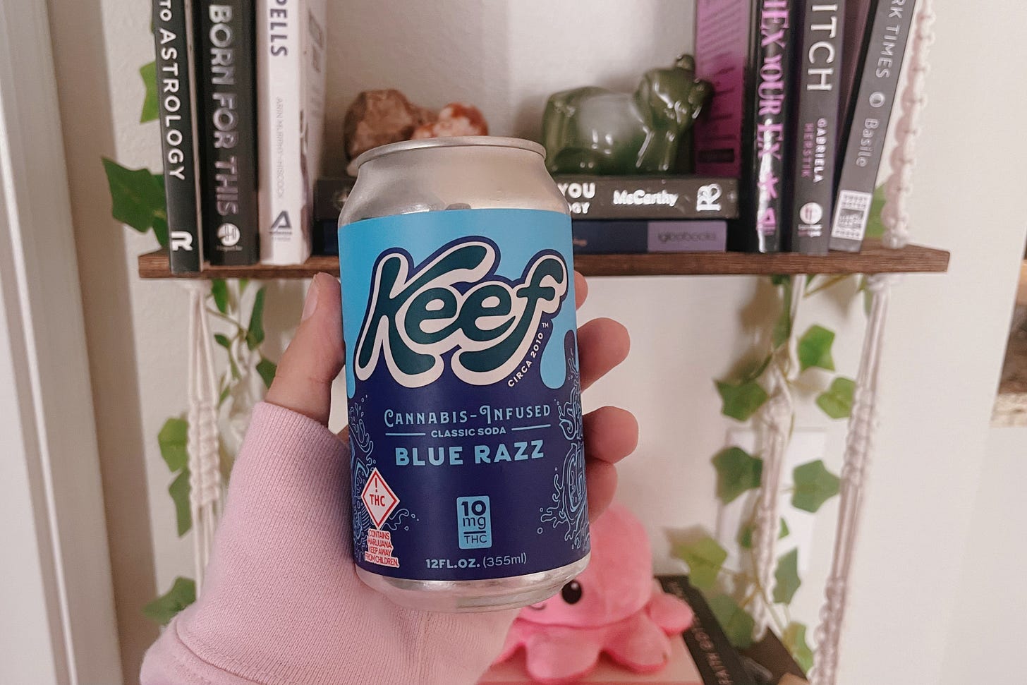 a canned drink, the label says "Keef cannabis-infused classic soda: Blue Razz", there is a shelf with some trinkets in the background