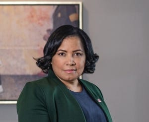 A photograph of a woman in a green coat and navy shirt sternly facing the camera