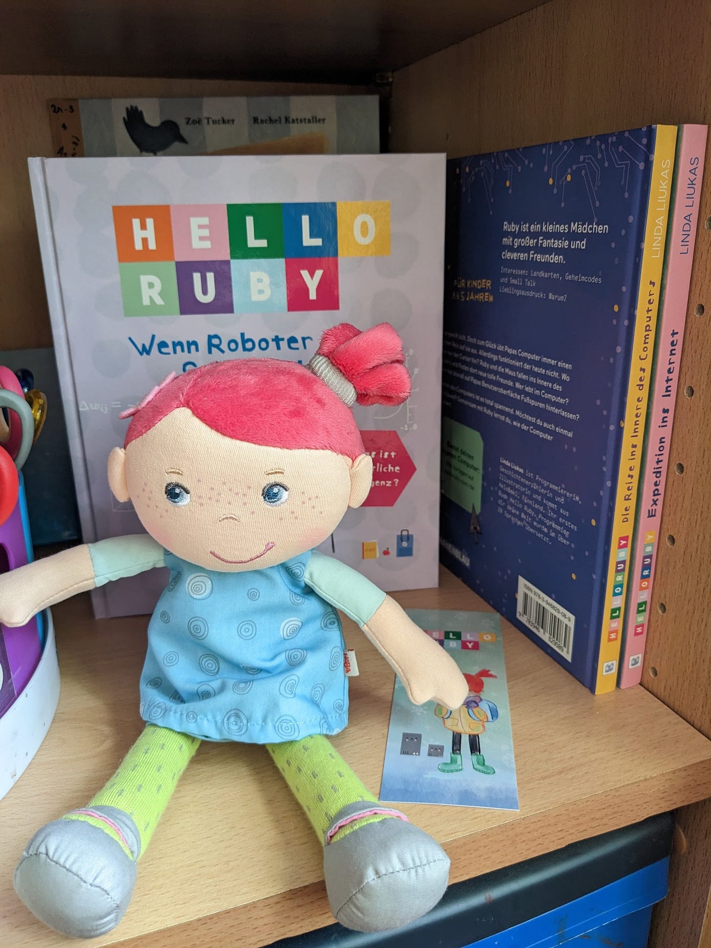 Eine Puppe mit roten Haaren, einem blauen Kleid und einer grünen Hose sitzt im Regal vor den Bücher "Hello Ruby" von Linda Liukas.