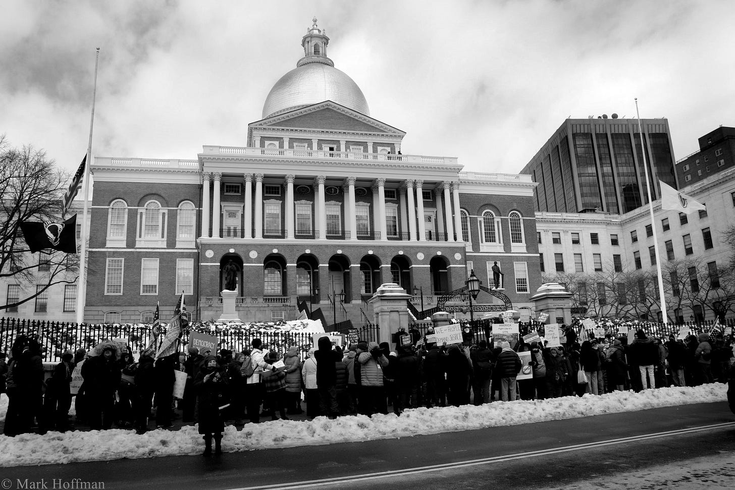 A crowd of protestors holding signs gather in front of the Massachusetts capitol building in the snow.