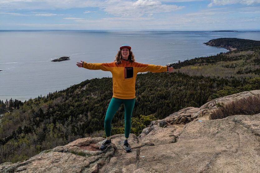 Haley, a young woman in a Cotopaxi sweatshirt and hat, stands on the summit of the Beehive Hike in Acadia National Park