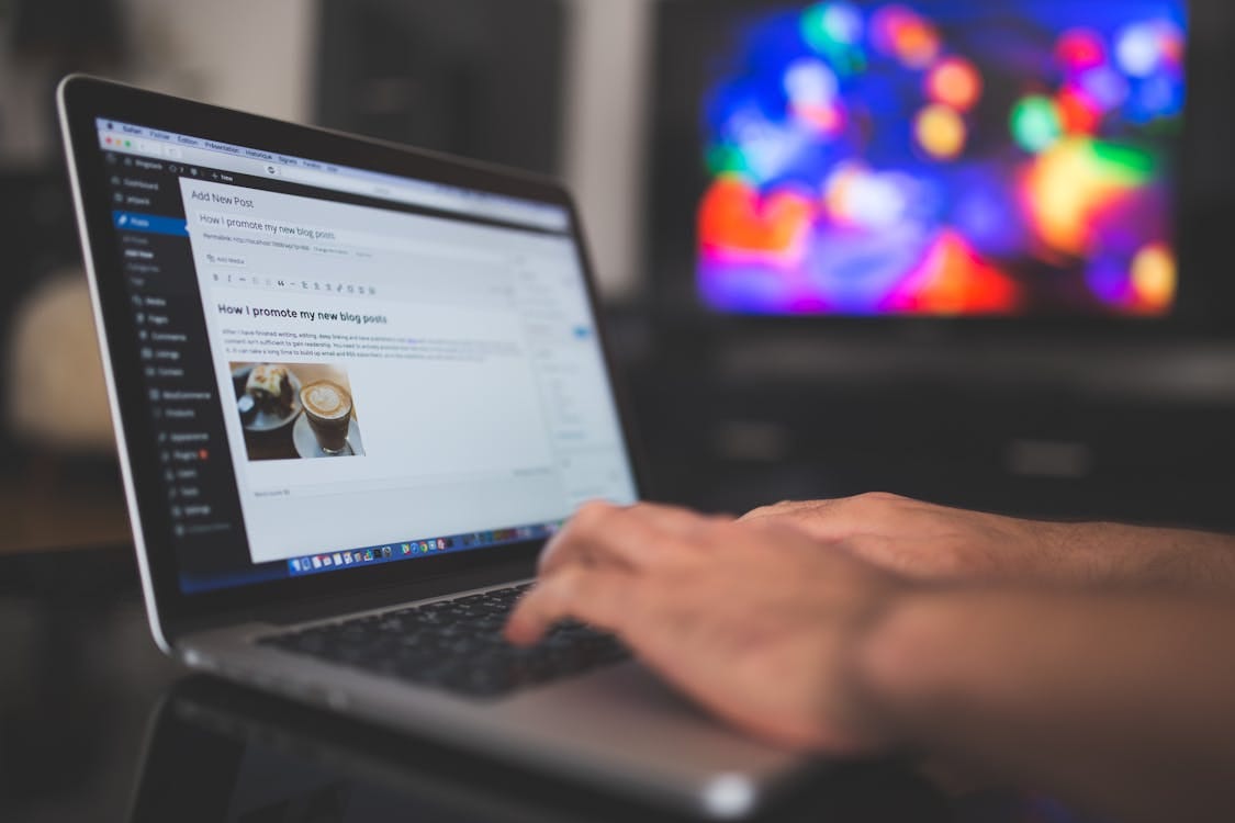 Free Hands typing on a laptop with a blog post visible, cozy indoor setting with colorful screen in background. Stock Photo