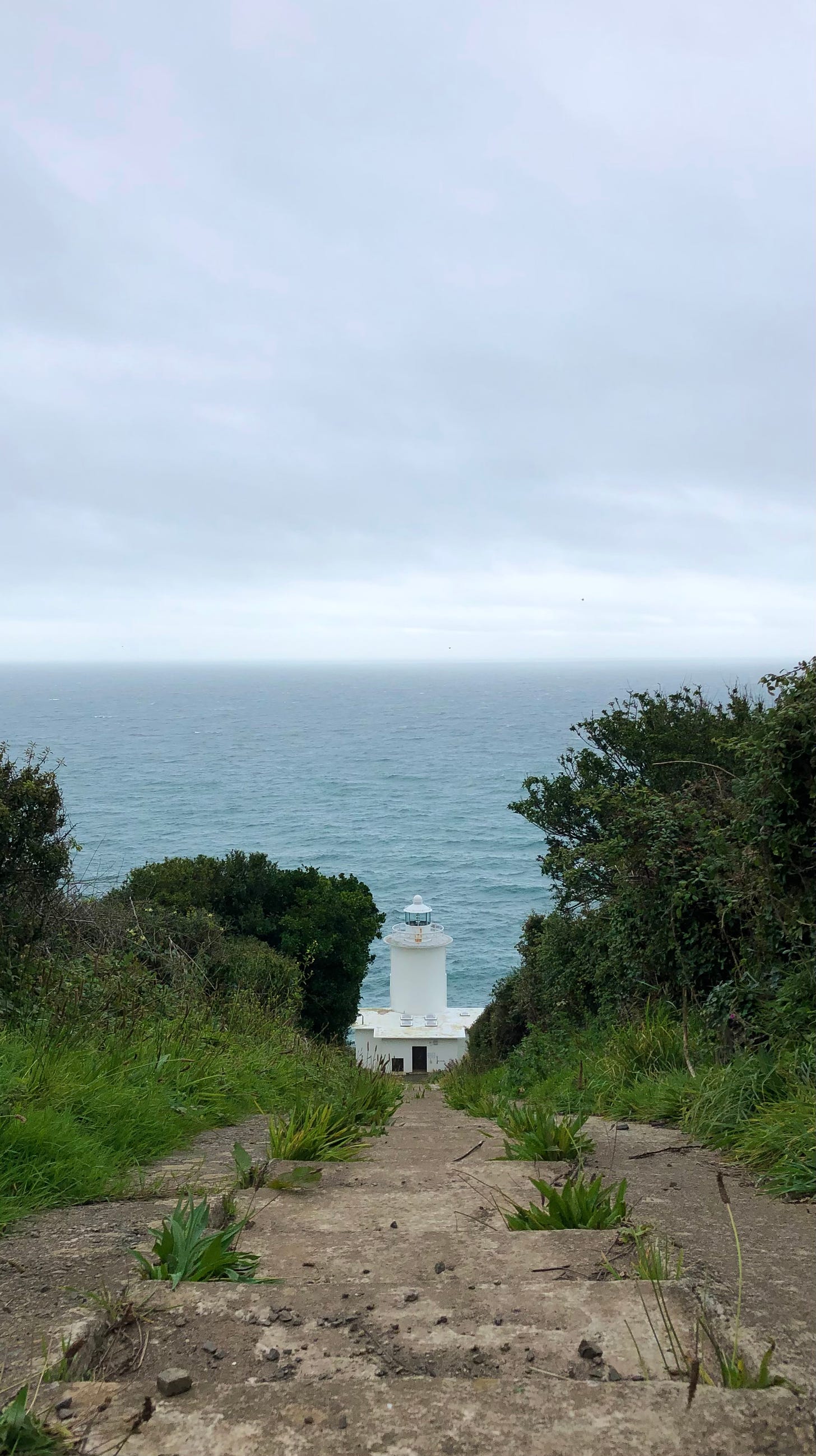 A photo looking down a steep set of stone steps to the white tower of a lighthouse with blue sea and light grey clouds beyond.