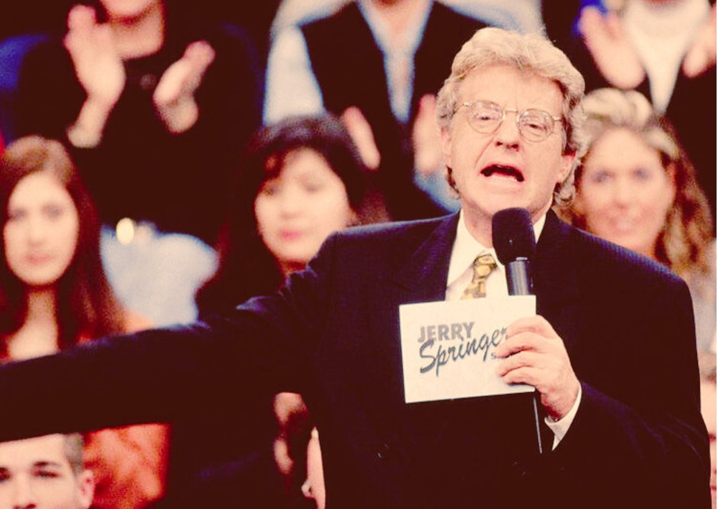 A picture from the Jerry Springer show; Springer in front of the audience, cards and microphone in hand, with a nostalgic filter applied.