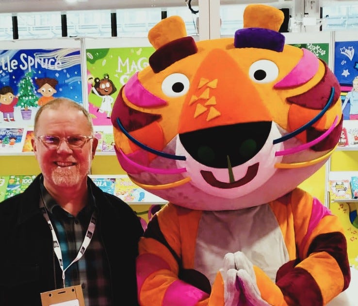 Author Troy Ford stands next to someone wearing a smiling tiger costume at the London Book Fair, 2024