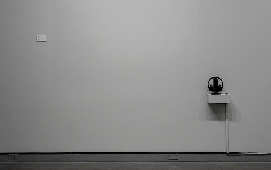 A photo of a white wall of a gallery shows wall text separate from a pair of headphones on a ledge.