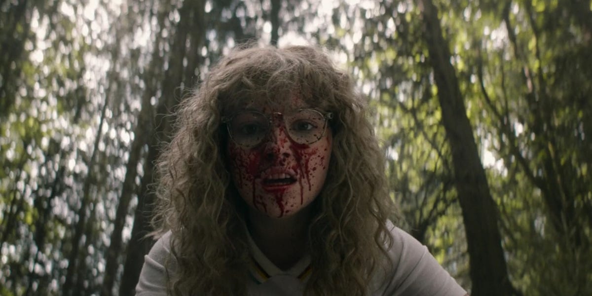 A young woman with blond curly hair and glasses stares at the camera with blood on her face