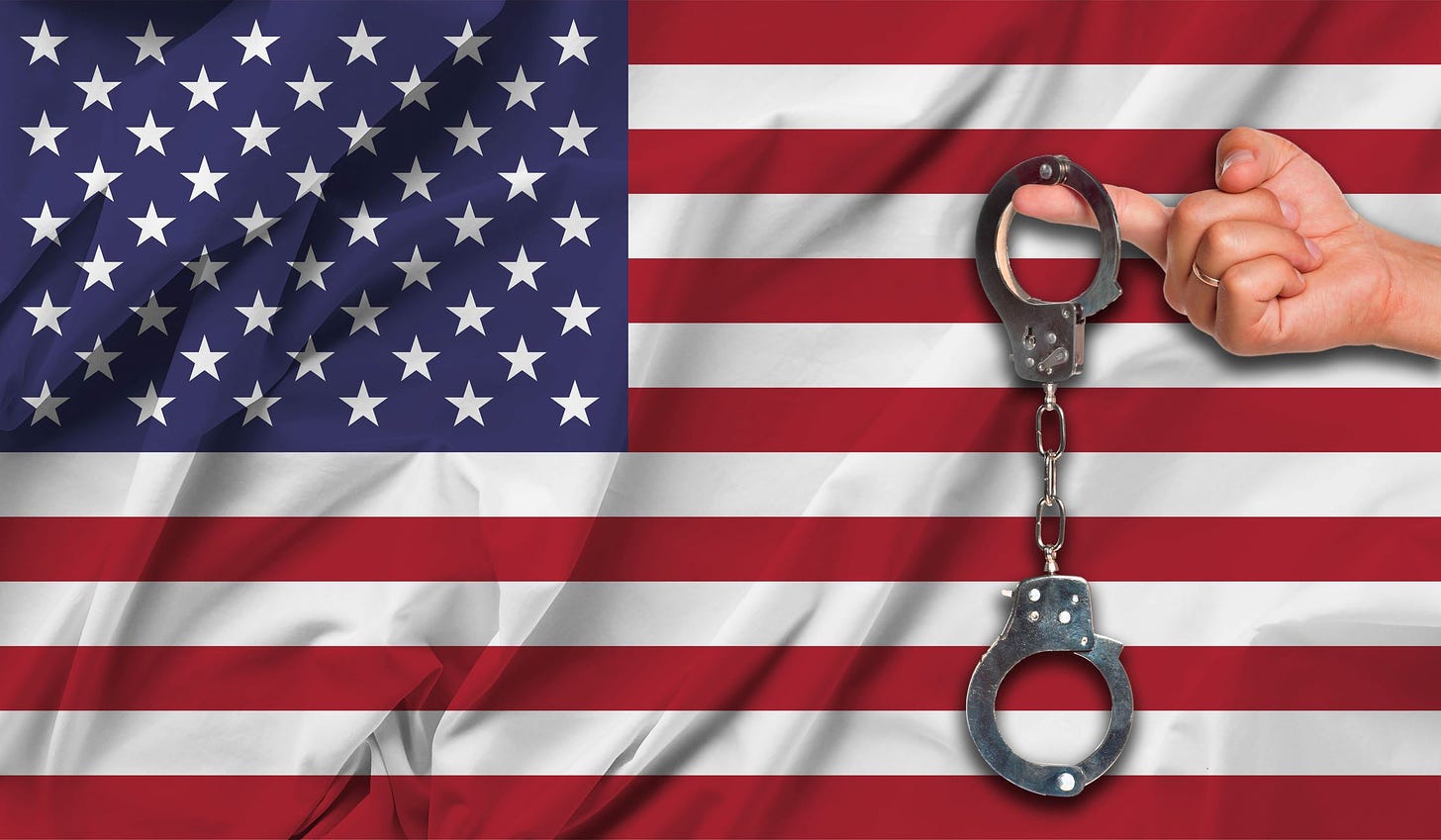 Photo of an American flag with a hand in front of it, holding a pair of handcuffs.