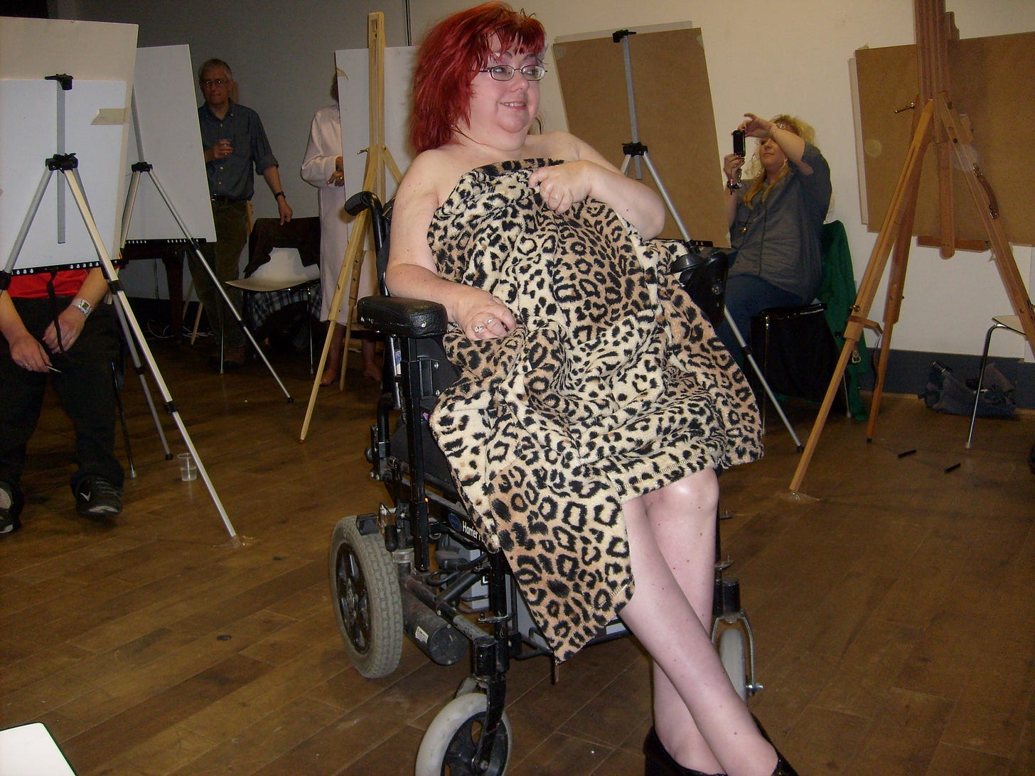 Penny sits in her wheelchair looking to one side. She is holding a loose leopard print fabric over her body, but exposes her legs to the knees and her shoulders are bare. Penny wears liked framed glasses and has deep red hair. She is in a room set up for art students who are busy behind their easels which stand on tripod frames. The scene is quite dark but Penny is smiling at her work as a model.