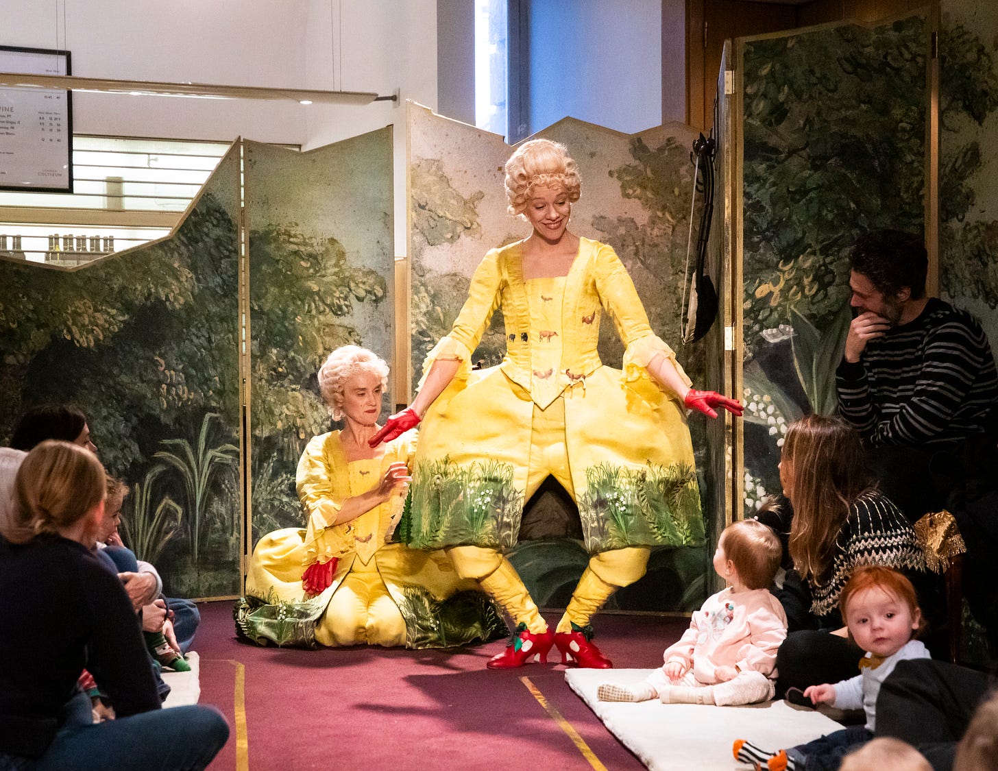 Two opera singers in yellow dresses and wigs perform in front of a wooden painted backdrop for babies and parents at ENO