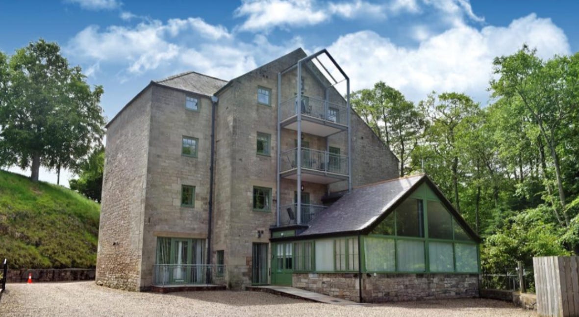 A massive four+ storey grey stone building with forest around and a glass conservatory at the front. There is a gravel driveway. 