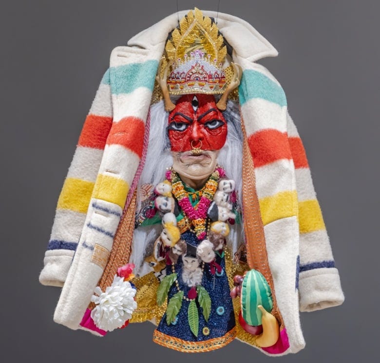 Closeup of a Hudson's Bay coat in the signature HBC colours of yellow, red, green, and off-white. The inner layer is intricately decorated with a red mask and gold crown, as well as plants and necklaces.