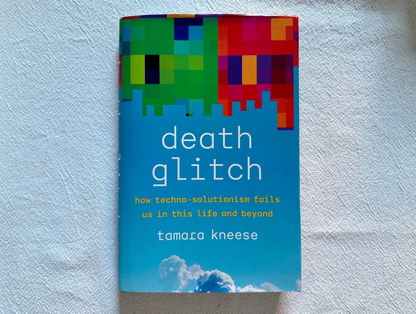 Image of the book "Death Glitch" by Tamara Kneese on a white tablecloth. The cover of the book is bright blue with a green and red pixelated pattern that appears to be falling from the top of the cover.