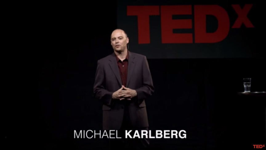 This color photograph shows Dr. Michael Karlberg standing on the TEDxInnsbruck stage giving his TED Talk "Beyond the Culture of Contest." Karlberg stands in front of the red TEDx logo, while his name (Michael Karlberg) appears in white letters beneath his image. This color photograph shows Dr. Michael Karlberg standing on the TEDxInnsbruck stage giving his TED Talk "Beyond the Culture of Contest." Karlberg stands in front of the red TEDx logo, while his name (Michael Karlberg) appears in white letters beneath his image.