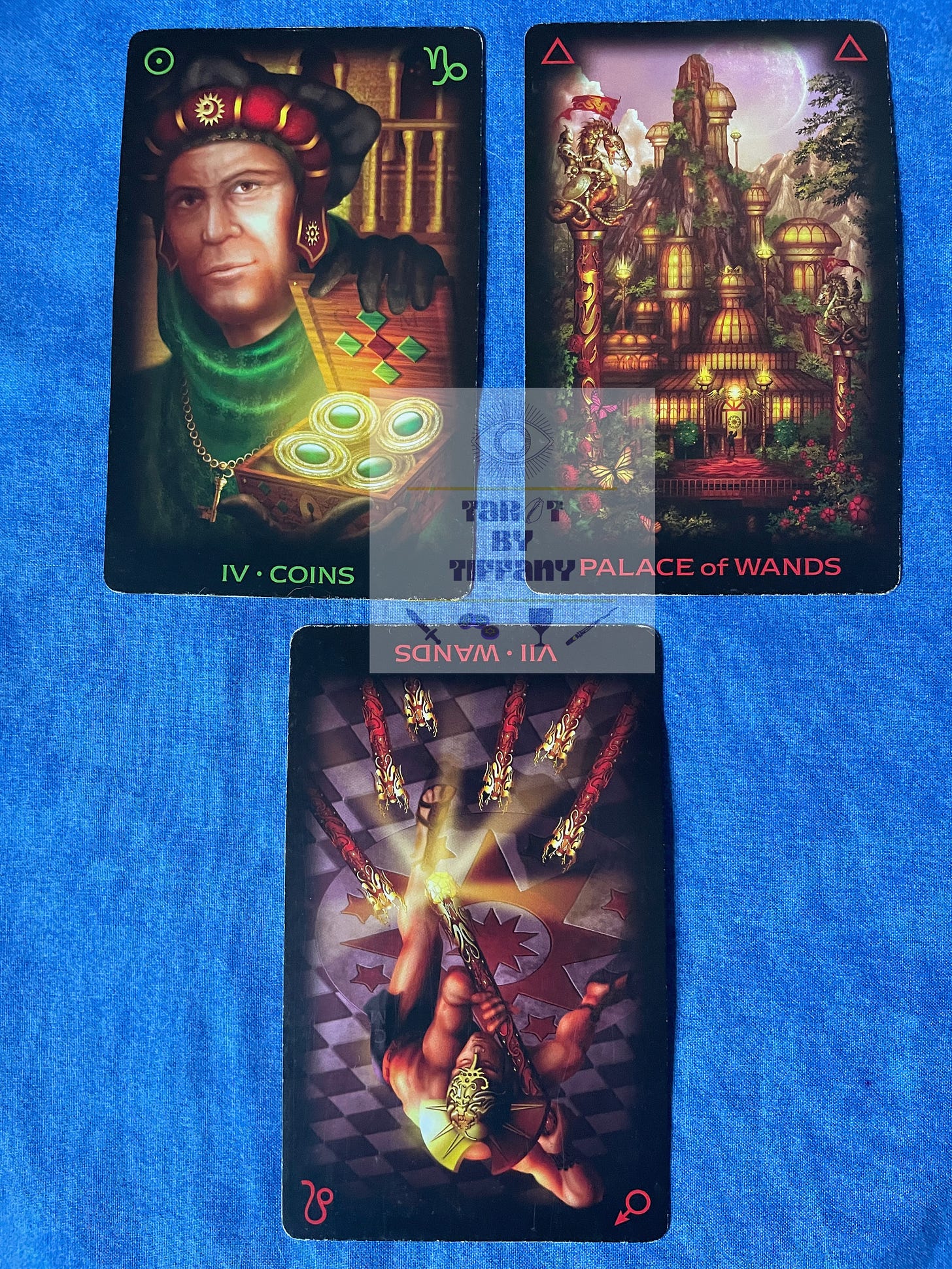 3-card reading with the Tarot of Dreams. The cards are laid against a cobalt blue cloth background. Top two cards (L-R): 4 of Coins, Palace of Wands. Bottom card: 7 of Wands reversed.