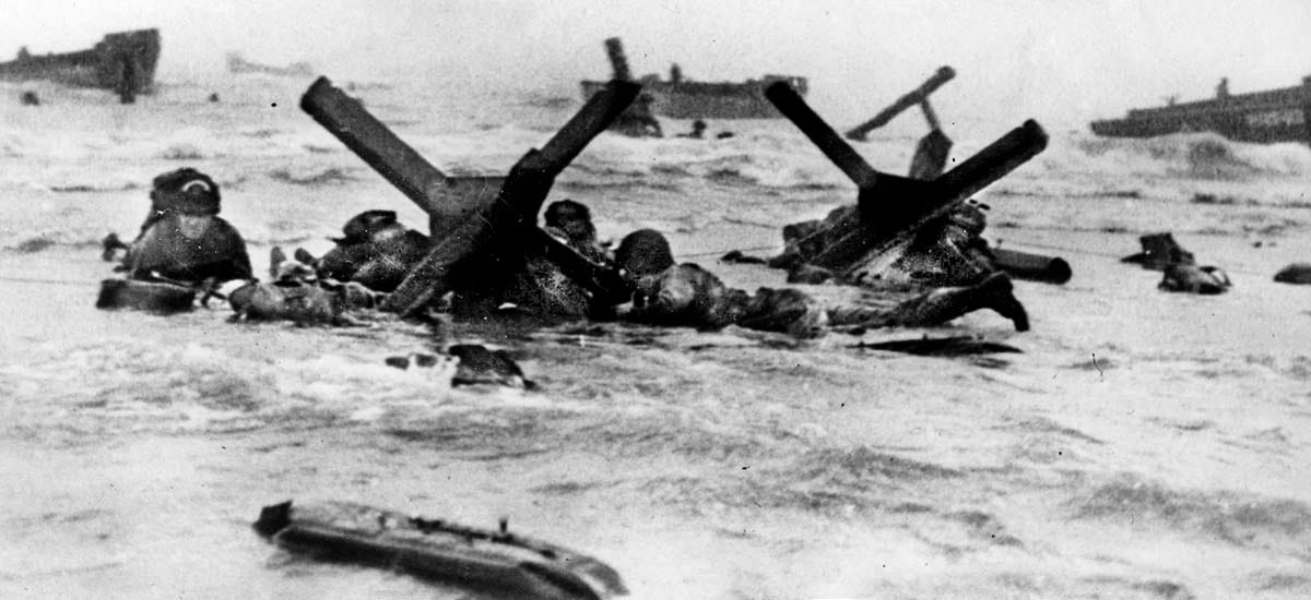 US Army soldiers in waist-deep ocean water crouch behind a steel-beamed barrier meant to impede beach landings