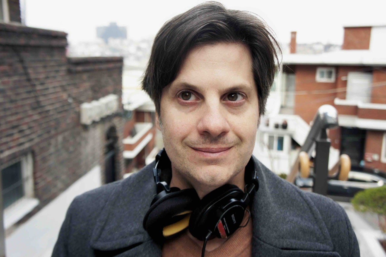 In this headshot, Jason, an American man with short black hair is wearing a grey coat over a brown jersey. His black headphones are around his neck and he is smiling into the camera.