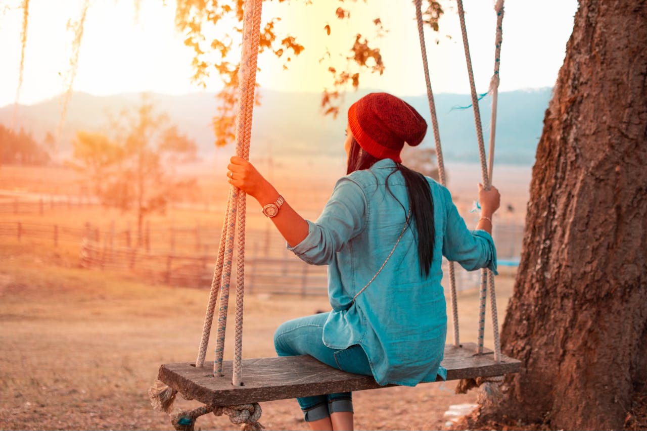 Woman sitting on a swing next to a tree with the sun setting in the distance.
