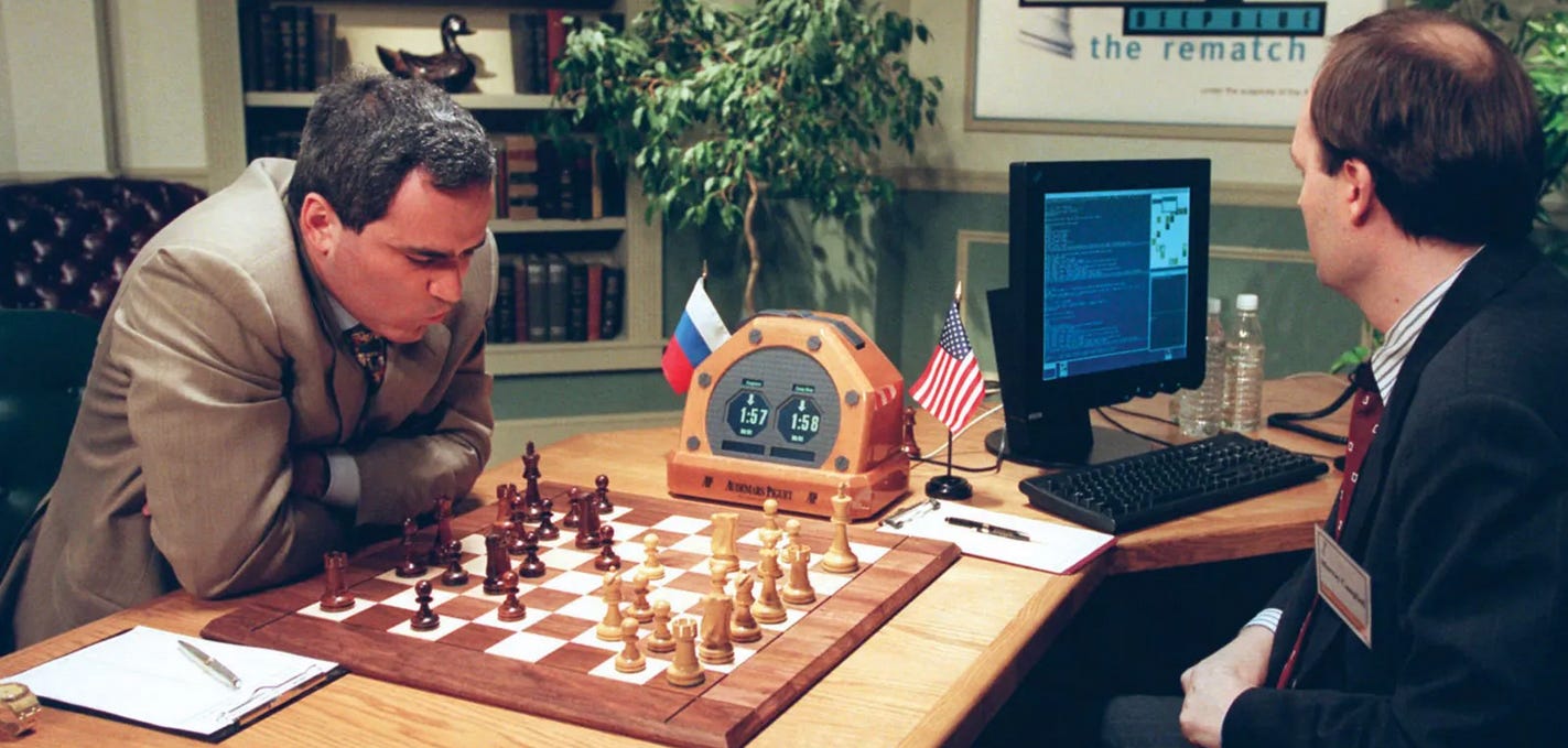 Foto da partida entre Garry Kasparov e o computador Deep Blue, controlado por um homem branco, calvo de terno e gravata.