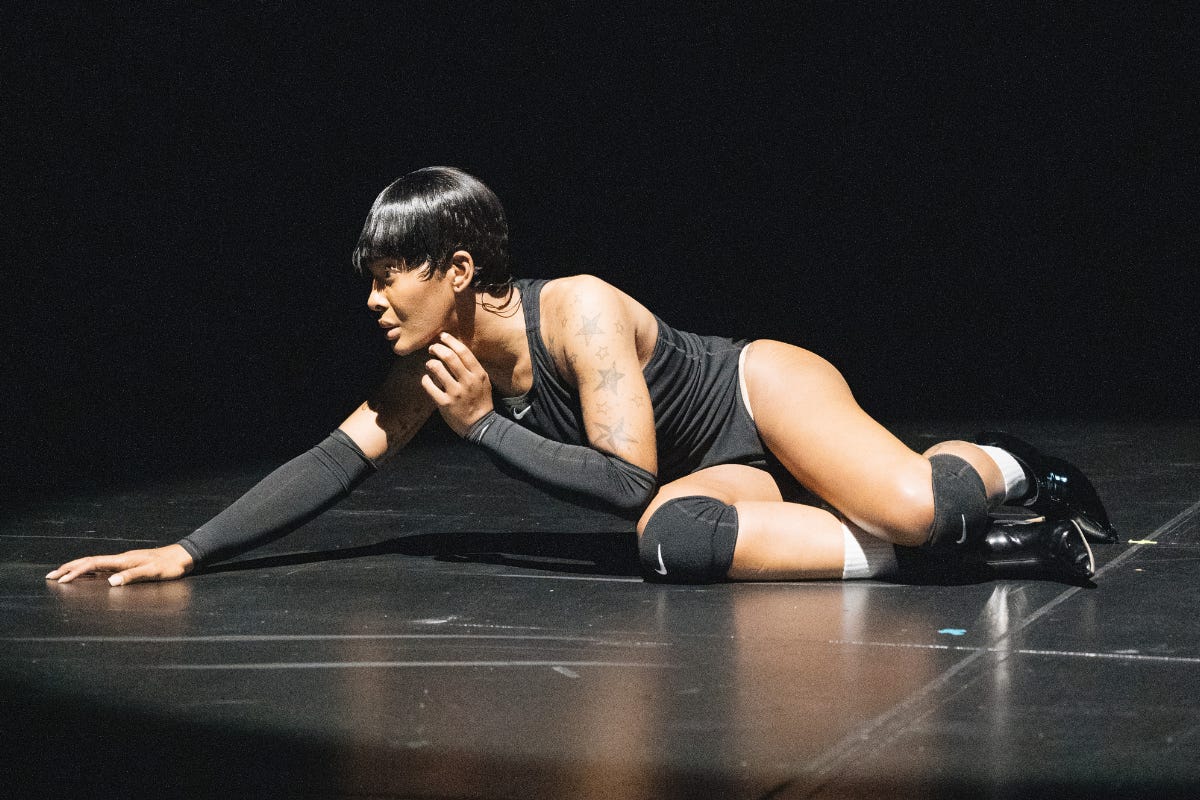 Ms. Boogie, a performer, is captured mid-performance lying on the stage floor. She wears a black sleeveless bodysuit, black knee pads, black gloves, and shiny black boots. Her short, sleek hairstyle and tattoos are highlighted under the dramatic stage lighting. Her pose, with one hand reaching forward and her face turned to the side, conveys intensity and focus against the dark background.