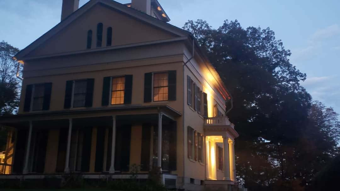 Dickinson's yellow house with green shutters in the evening. The house Federal Architecture style and is shadowed and the lights are on in the far right two windows, which are her room. Below a porch with four columns. To the right the porch light illuminates the front of the house. Behind the house the full dark trees and the dim blue sky. 