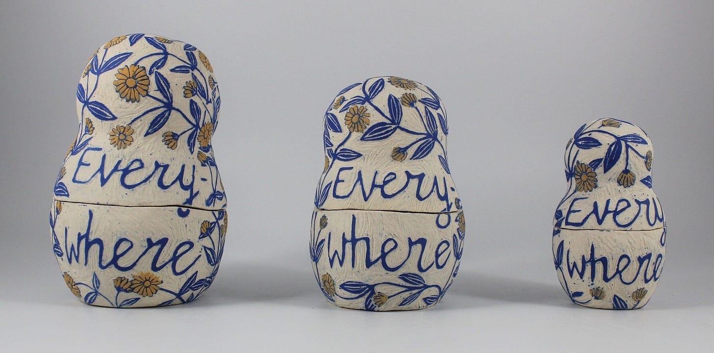  Three hand-painted matryoshka (nesting) dolls, arranged from largest to smallest, each decorated with blue leaves, yellow flowers, and the word "Everywhere" written across their midsections in cursive. The background is plain light grey, and the dolls have a textured, off-white surface.