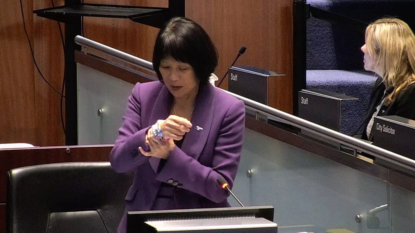 Mayor Olivia Chow, in a purple suit, stands at her lectern in the Council chamber, holding up bracelets on her right wrist