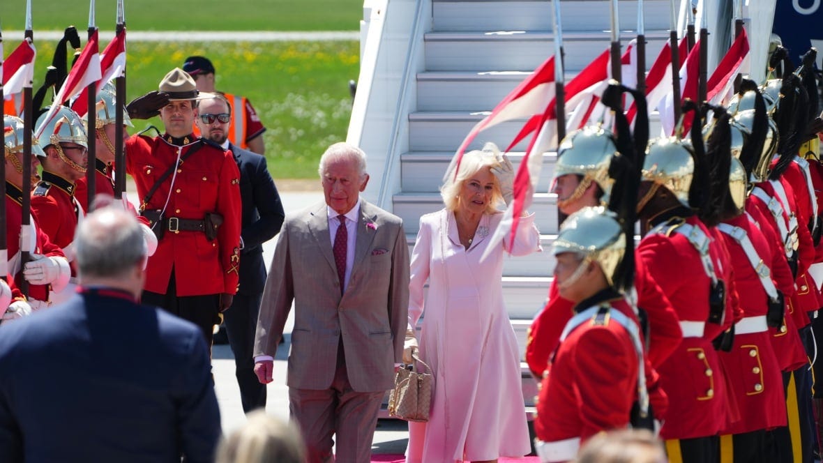 A man in a beige suit and a woman in a pale pink dress walk on a red carpet near plane stairs and guards.
