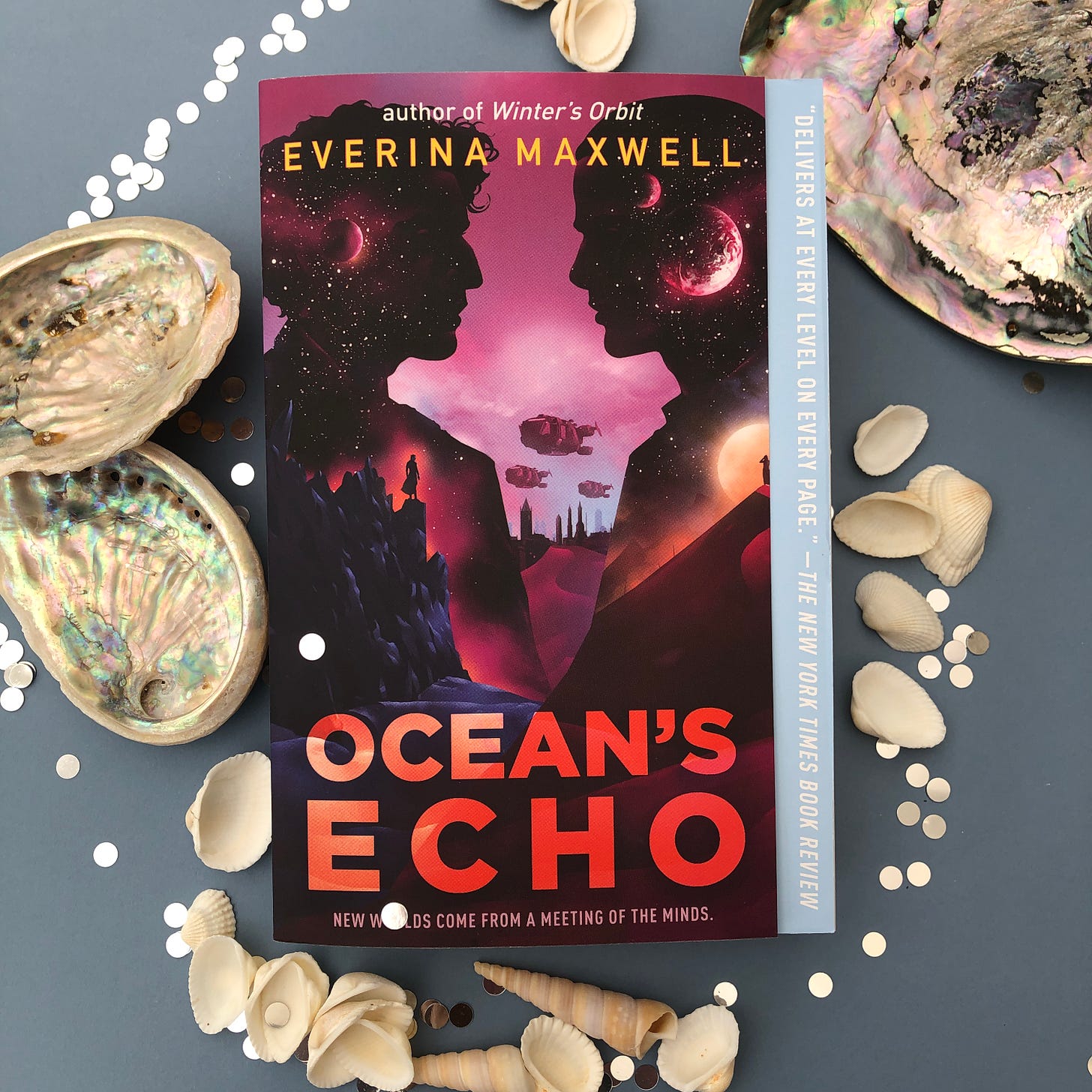 The paperback of Ocean's Echo surrounded by shells. The cover features two men facing each other over a purple and red futuristic city landscape.