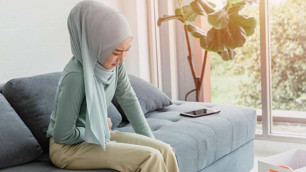 A young Arabic woman sitting on a sofa, clutching her abdomen in pain