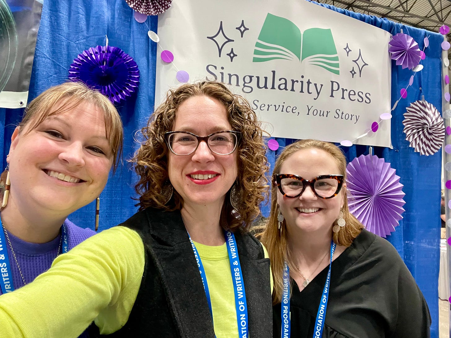A selfie of three white women, one with hair pulled back and dangling earrings, one with long strawberry blond hair and dangle earrings and big glasses, one in the middle (me) in a. yellow sweater and gray vest. Behind us is a sign that reads "Singularity Press" with purple accordion fans dangling around it.