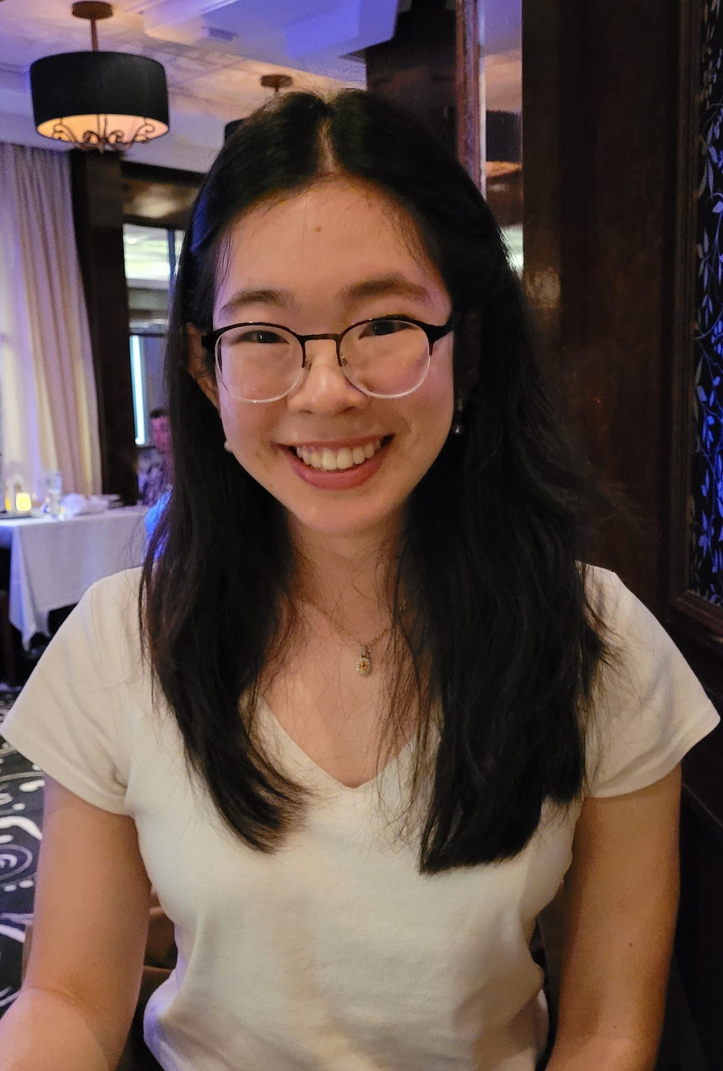 Smiling young woman with glasses and long black hair, wearing a white T-shirt.