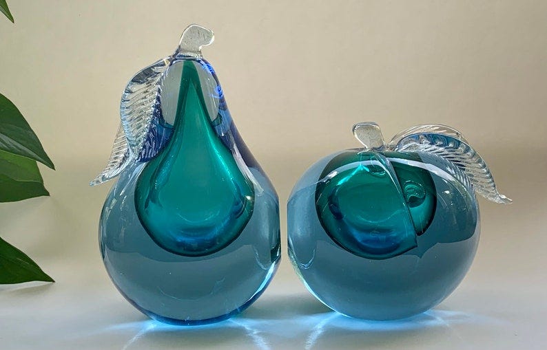 May include: Two glass sculptures, one shaped like a pear and the other shaped like an apple. Both sculptures are made of clear glass with a blue tint and have a teal blue interior. The pear has a clear glass stem and leaf. The apple has a clear glass stem and leaf.